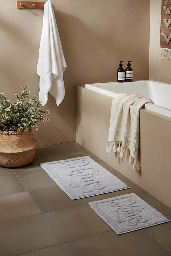 Hellgraues, geprägtes 2-teiliges Bad- und WC-Matten-Set aus Baumwolle mit dem Aufdruck „Home“.