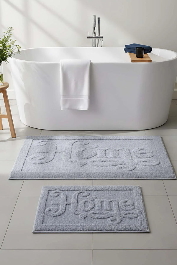 Hellgraues, geprägtes 2-teiliges Bad- und WC-Matten-Set aus Baumwolle mit dem Aufdruck „Home“.